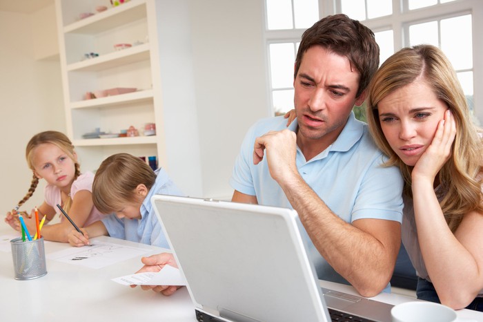 زوجان قلقان بشكل واضح يقرؤون موادًا على جهاز كمبيوتر محمول ، ويجلس أطفالهم في الخلفية.