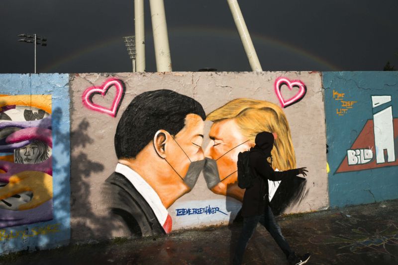 رجل يمشي أمام كتابات على الجدران تصور الرئيس الأمريكي ترامب ، على اليمين ، والرئيس الصيني شي جين بينغ يقبلان بعضهما البعض بأقنعة للوجه ، معروضة على جدار في الحديقة العامة Mauerpark في برلين ، ألمانيا ، الأربعاء ، 29 أبريل ، 2020. (AP Photo / ماركوس شرايبر)